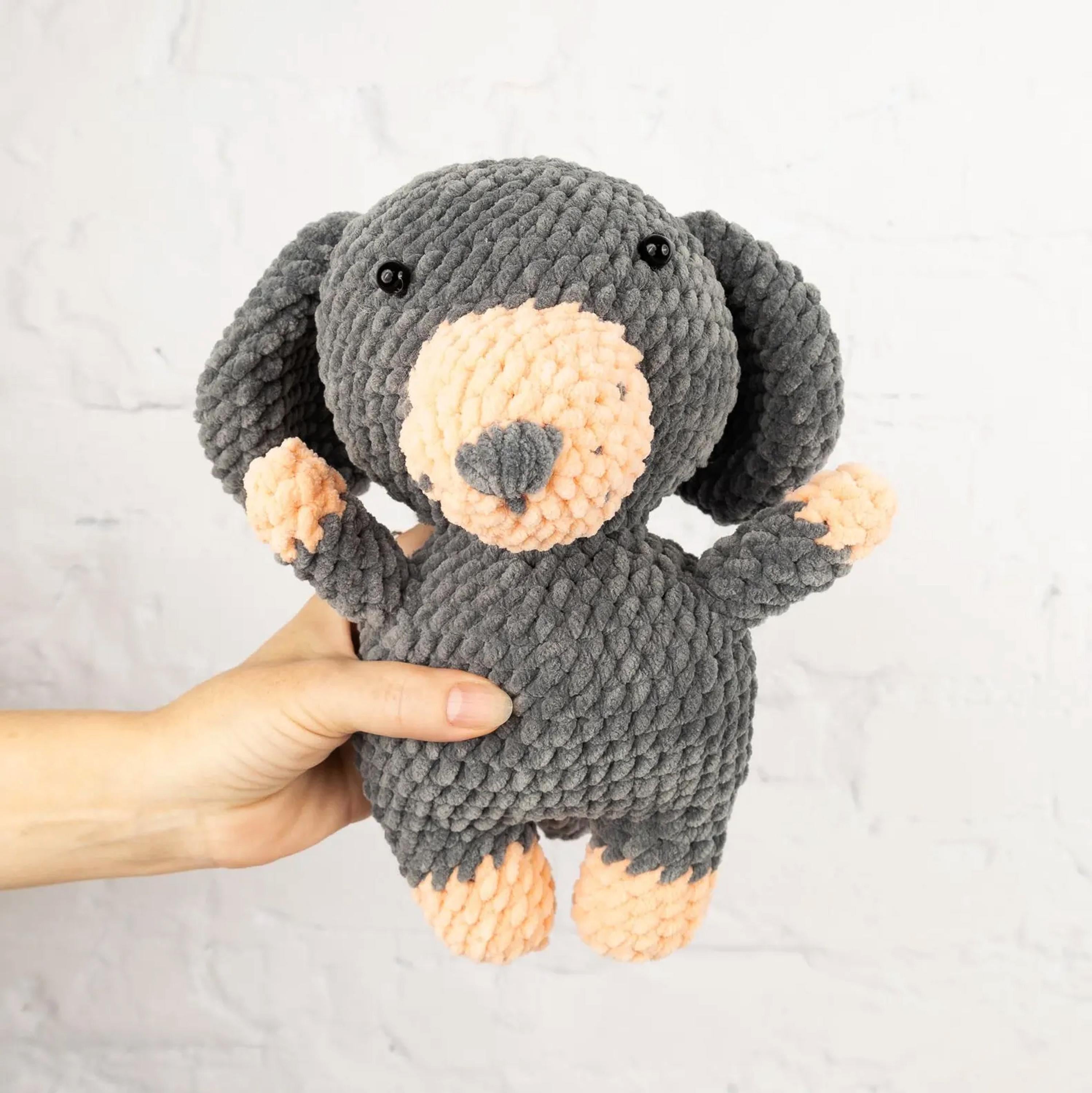 A hand is holding a gray and peach-colored stuffed animal, which appears to be a small dog or puppy.